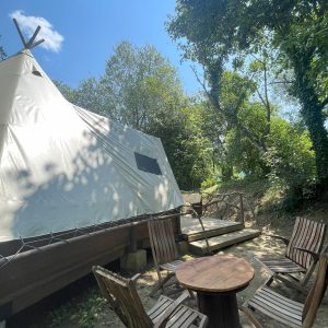 Tipi en Basse-Normandie, entouré de verdure, avec chaises en bois et table.