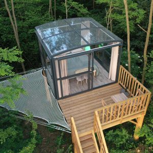 Cabane en verre perchée dans les arbres, avec terrasse en bois et vue sur la forêt.