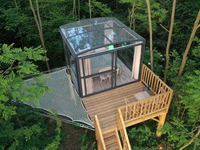 Cabane en verre perchée dans les arbres, avec terrasse en bois et vue sur la forêt.