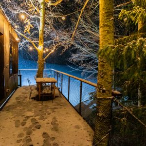 Cabane suspendue en bois, illuminée, avec une terrasse surplombant la neige.