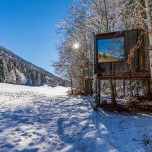 Cabane perchée en bois, entourée de neige et de montagnes sous un ciel bleu.