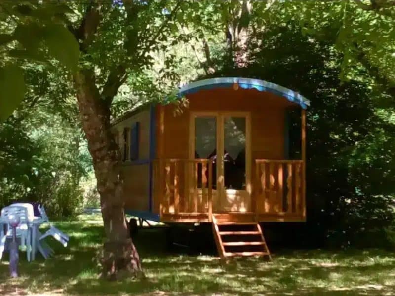 Chalet en bois avec terrasse, entouré darbres verdoyants et mobilier de jardin.