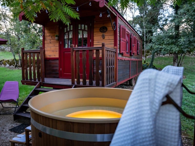 Chalet en bois avec terrasse, jacuzzi en bois et chaises longues dans un cadre verdoyant.