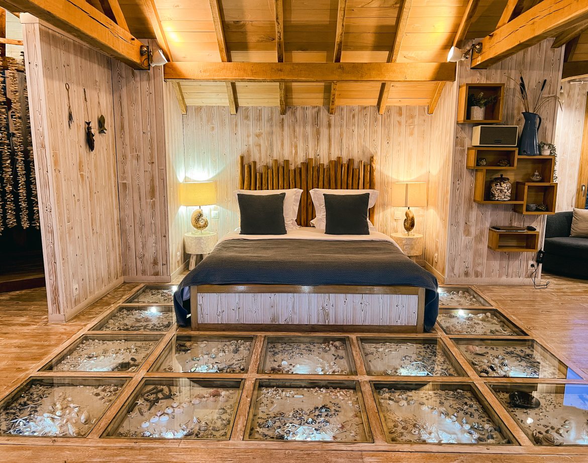 Chalet en bois avec un lit confortable et un sol en verre révélant des éléments naturels.