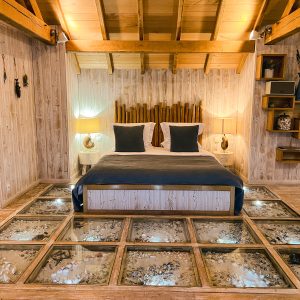 Chalet en bois avec un lit confortable et un sol en verre révélant des éléments naturels.