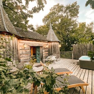 Cabane en bois avec tourelles, terrasse en bois et jacuzzi entouré de verdure.
