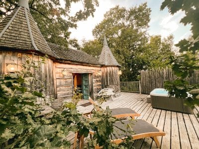 Cabane en bois avec tourelles, terrasse en bois et jacuzzi entouré de verdure.