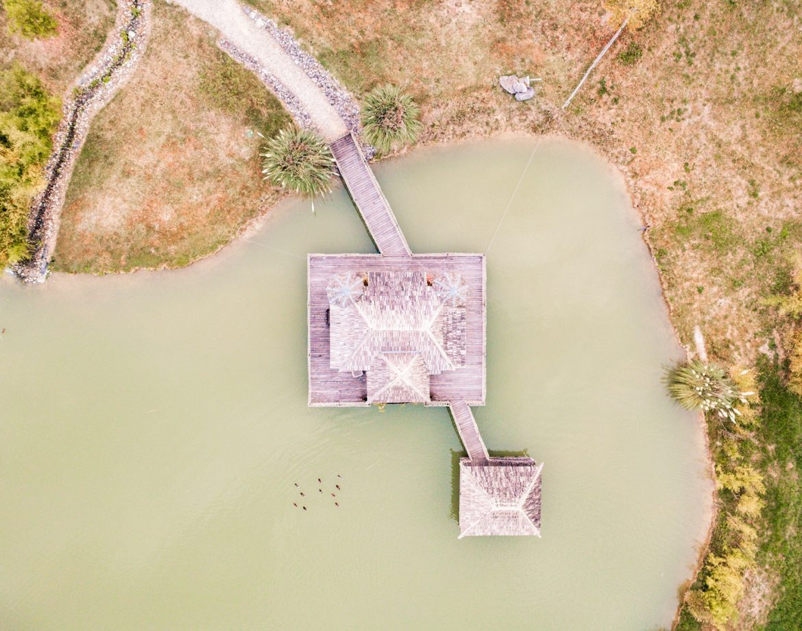 Cabane flottante sur un étang, entourée de verdure et accessible par un pont.