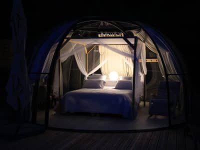 Hébergement insolite en bulle transparente, lit romantique éclairé par une douce lumière.