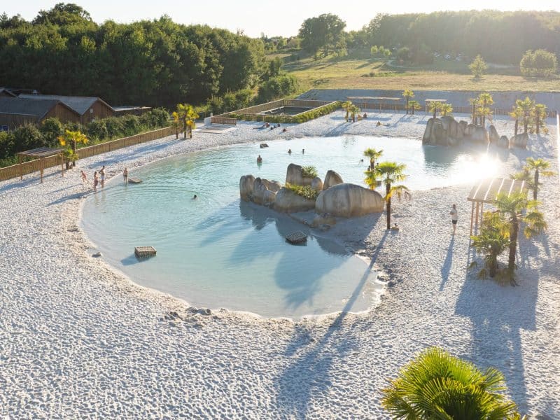 Hébergement touristique avec piscine naturelle et sable blanc, entouré de palmiers.