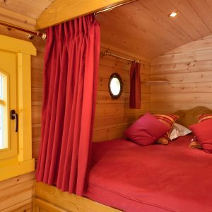 Cabane en bois avec un lit douillet, rideaux rouges et fenêtre ronde.