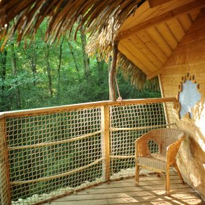 Cabane perchée en bois avec balcon en bois et fauteuil en osier, entourée de verdure.