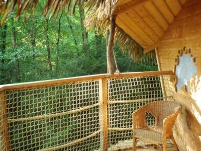 Cabane perchée en bois avec balcon en bois et fauteuil en osier, entourée de verdure.
