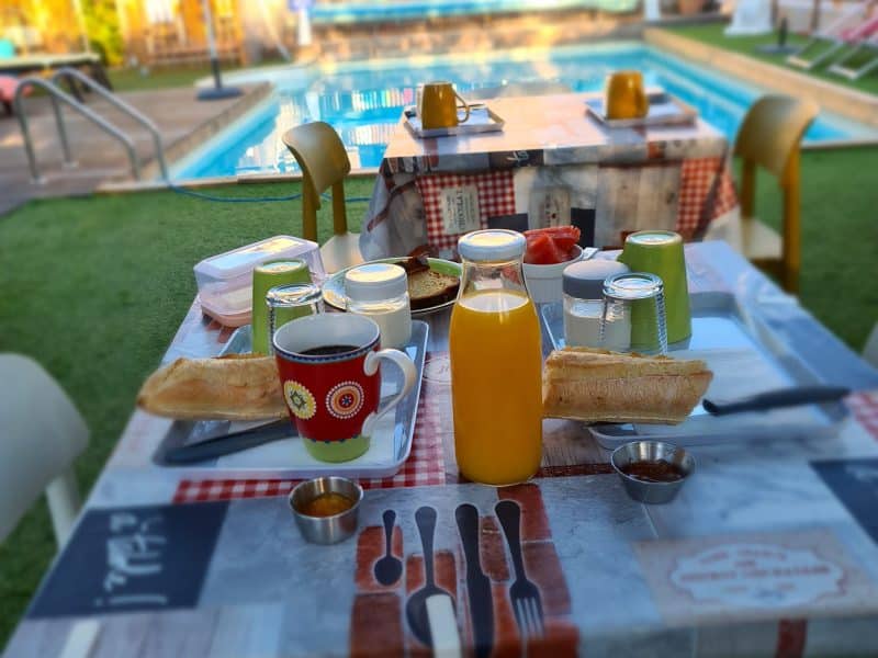 Table de petit-déjeuner en plein air près dune piscine, avec jus et viennoiseries.