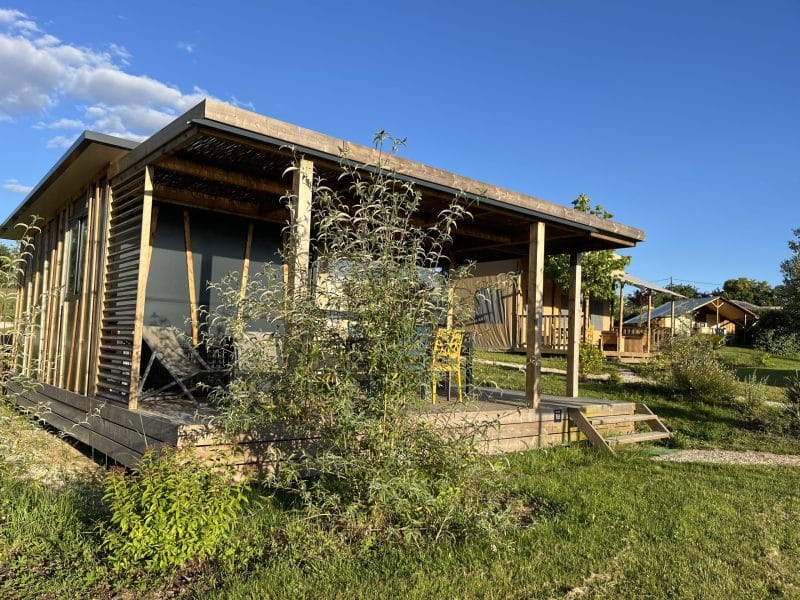 Cabane en bois avec terrasse, entourée de verdure et sous un ciel bleu.
