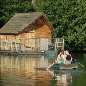 Cabane en bois au bord de leau, avec des visiteurs en barque. Nature verdoyante.