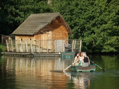 Cabane en bois au bord de leau, avec des visiteurs en barque. Nature verdoyante.