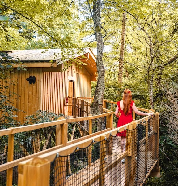 Cabane perchée dans les arbres, entourée de verdure, avec une passerelle en bois.