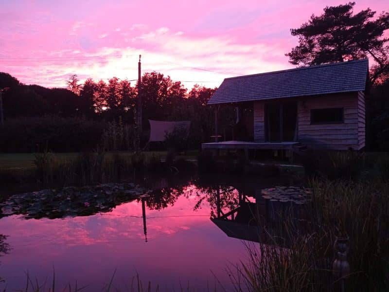 Chalet en bois au bord dun étang, reflet du ciel rose au coucher du soleil.
