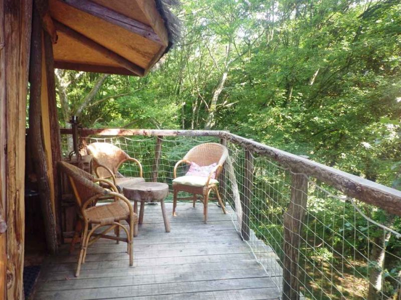 Cabane dans les arbres avec terrasse en bois et chaises en rotin, entourée de verdure.