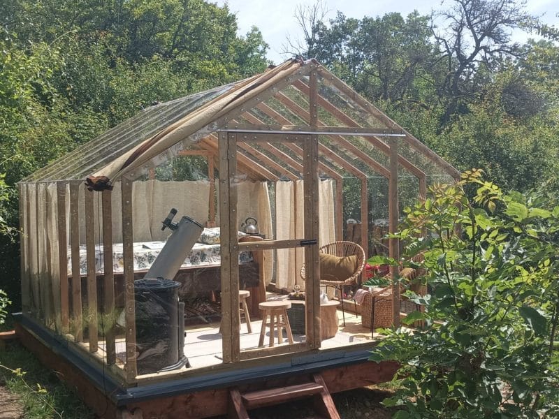 Cabane en bois avec grandes fenêtres, entourée de verdure et dun télescope.