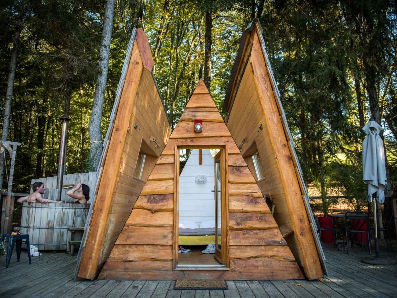 Cabane en bois en forme de A, entourée darbres, avec terrasse accueillante.