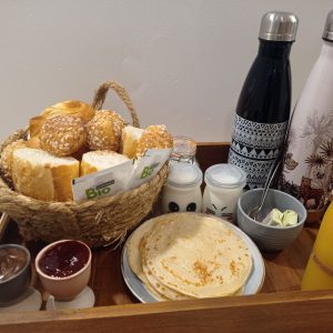 Panier de petit-déjeuner avec pains, crêpes et boissons dans un hébergement insolite.