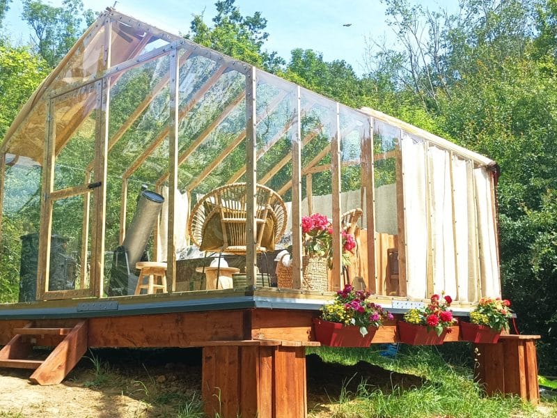 Cabane en verre entourée de verdure, avec des fleurs colorées sur la terrasse.