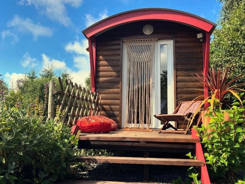Charmante roulotte en bois avec terrasse, entourée de verdure et dun ciel bleu.