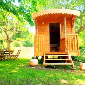 Charmante roulotte en bois, entourée de verdure, avec terrasse et mobilier de jardin.