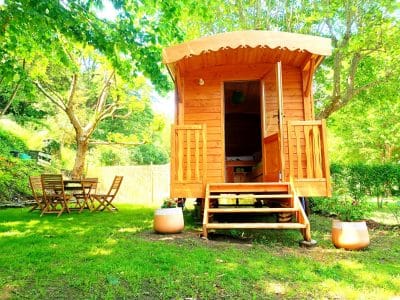 Charmante roulotte en bois, entourée de verdure, avec terrasse et mobilier de jardin.