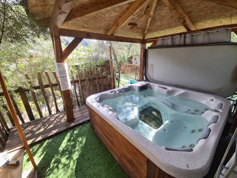 Hébergement en chalet avec jacuzzi privé sous un abri en bois, entouré de verdure.