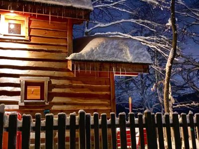 Cabane en bois sous la neige, éclairée par une douce lumière nocturne.