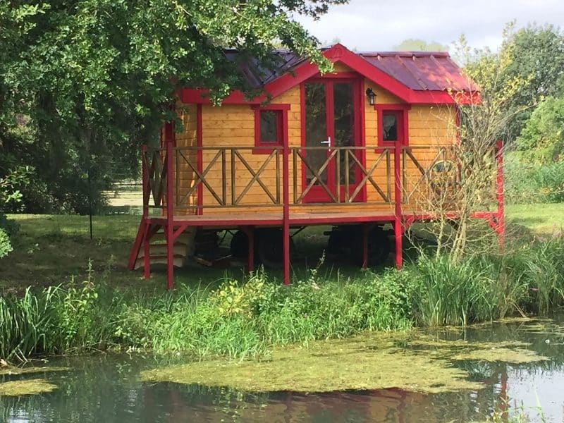 Cabane en bois sur pilotis, avec balcon rouge, au bord dun étang verdoyant.