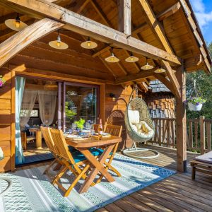 Cabane en bois avec terrasse ensoleillée et chaises suspendues, idéale pour se détendre.