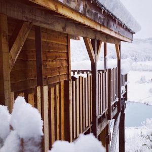 Cabane en bois perchée, entourée de neige, vue sur un lac paisible.