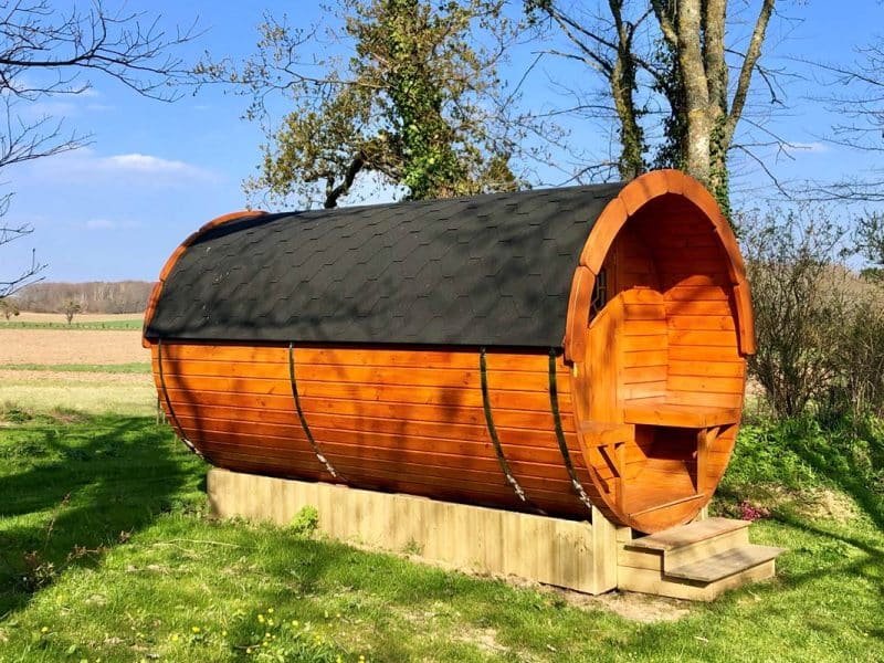 Cabane en bois en forme de tonneau, entourée de verdure et de champs.