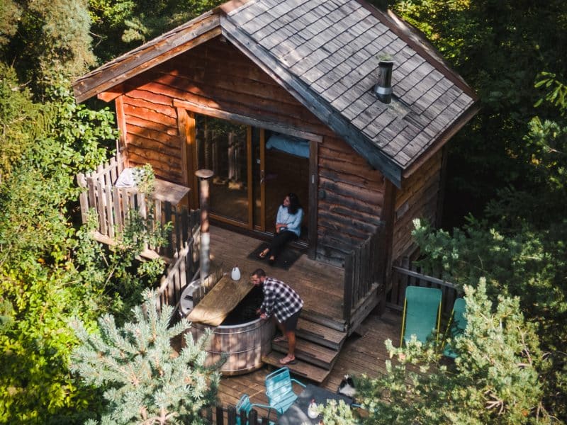 Cabane en bois entourée darbres, avec une terrasse accueillante et un jacuzzi.