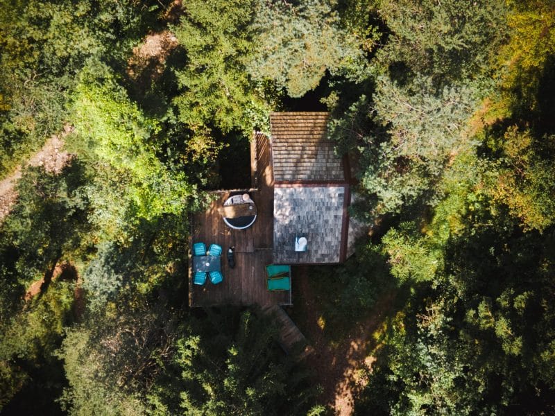Cabane en bois entourée de verdure, avec une terrasse et un jacuzzi.