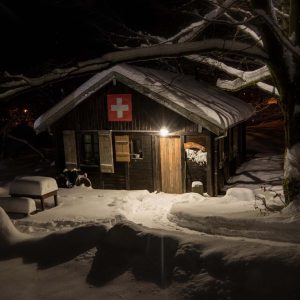 Cabane suisse en bois, illuminée, entourée de neige et de branches enneigées.