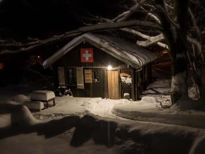Cabane suisse en bois, illuminée, entourée de neige et de branches enneigées.