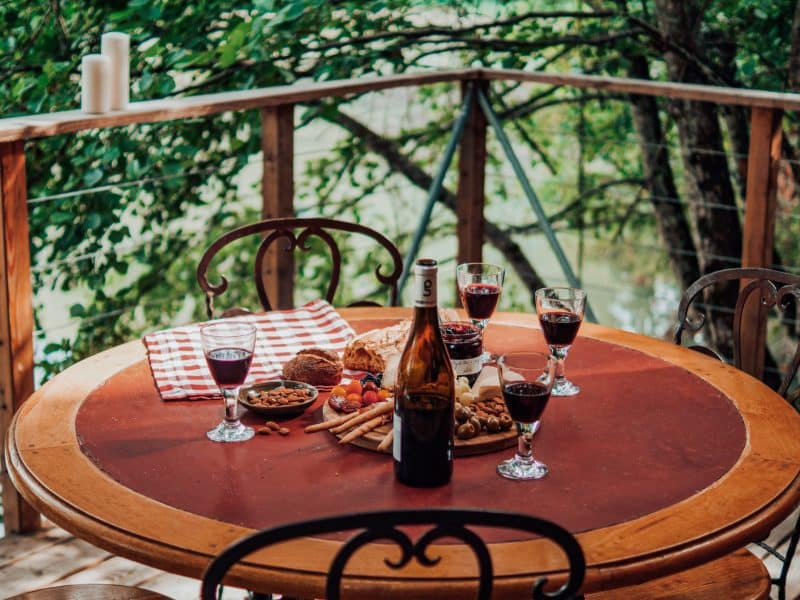Table en bois sur une terrasse, avec vin et snacks, entourée de verdure.