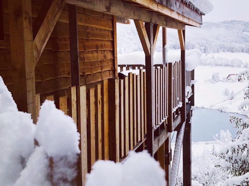 Chalet en bois enneigé avec balcon, offrant une vue sur un paysage hivernal.