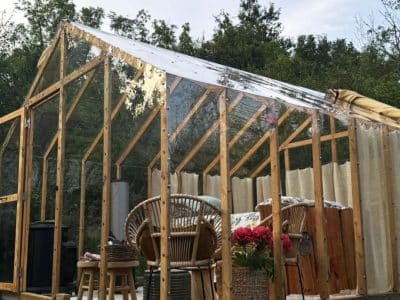 Cabane en verre dans la nature, avec mobilier en rotin et fleurs colorées.