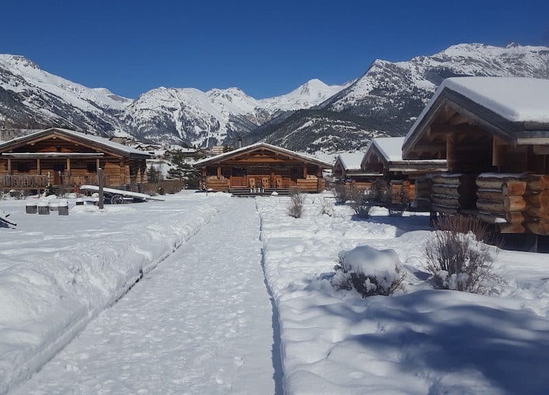 Chalets en bois dans un paysage enneigé avec montagnes majestueuses en arrière-plan.