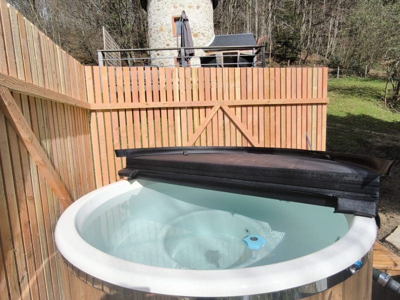 Gîte en pierre avec jacuzzi en bois, entouré de verdure et dune clôture en bois.