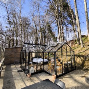 Hébergement insolite en serre vitrée, entouré darbres, avec terrasse en bois.