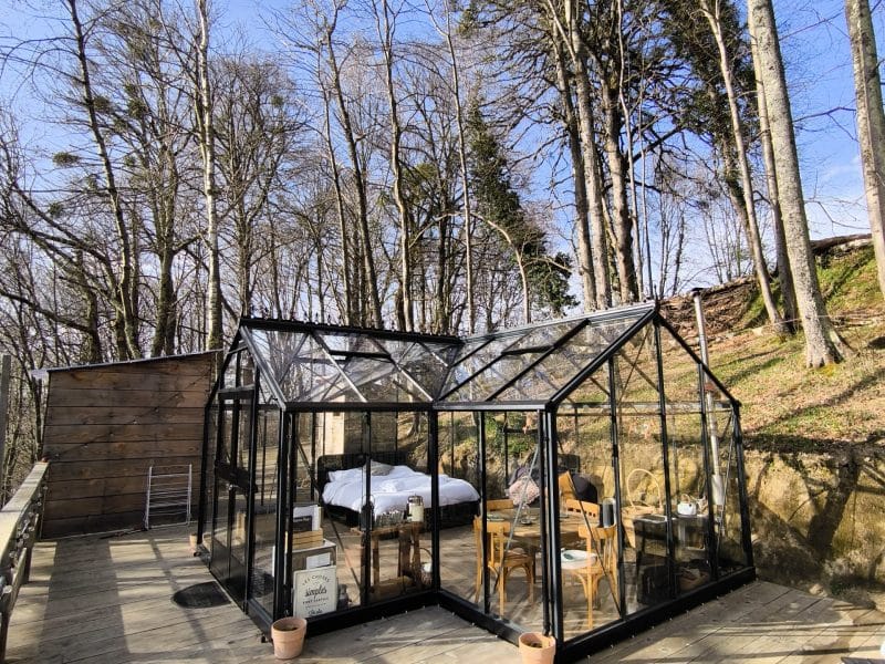 Hébergement insolite : une serre en verre entourée darbres, avec mobilier en bois.