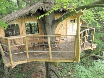 Cabane dans les arbres en bois, avec terrasse suspendue et vue sur la forêt verdoyante.