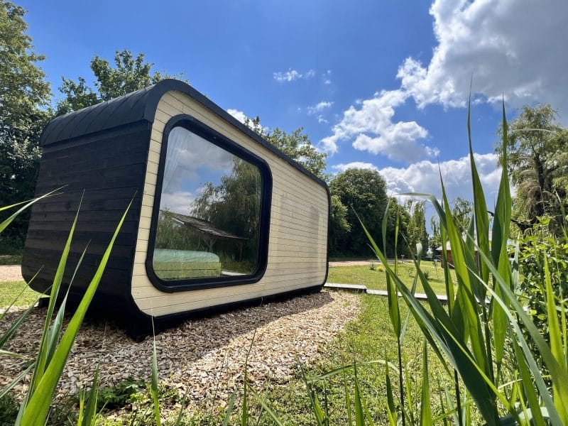 Cabane moderne en bois, avec de grandes fenêtres, entourée de verdure et sous un ciel bleu.